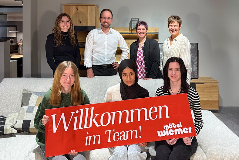 8 Personen, davon sitzen 3 auf einem Sofa mit einem Schild "Wilkommen im Team Möbel Wiemer"