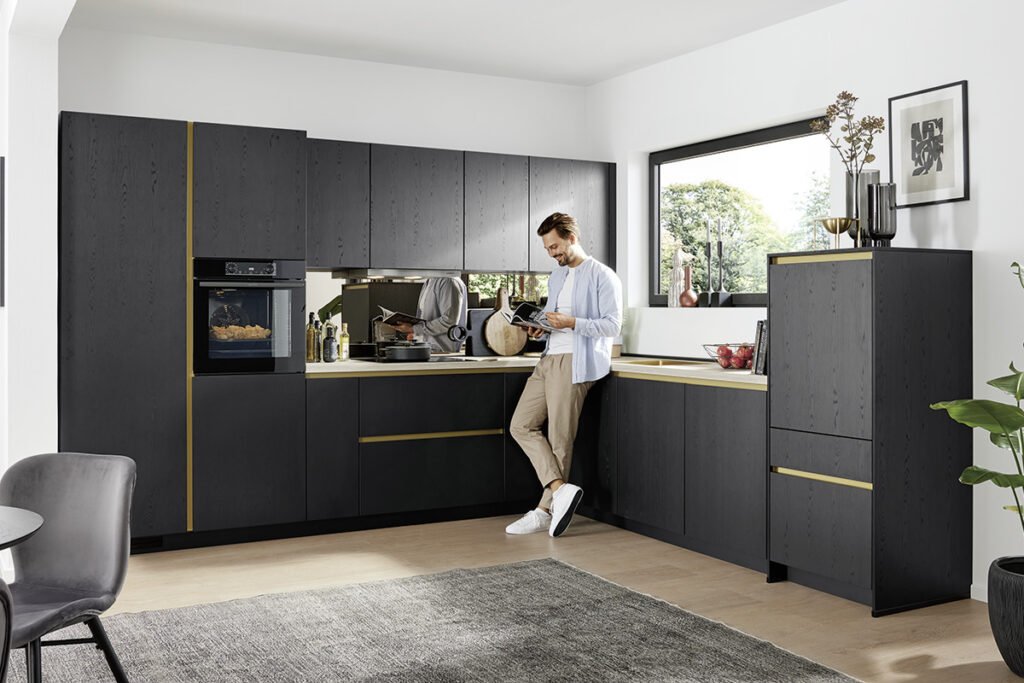 Front Eiche Nero Nachbildung, Arbeitsplatte Travertin Sandgrau Nachbildung, Nischenverkleidung Spiegel bronzefarbig, Griffmulden gelbgoldfarbig. Mit Elektrogeräten von Gorenje und goldfarbiger Einbauspüle. Ohne Deko. 